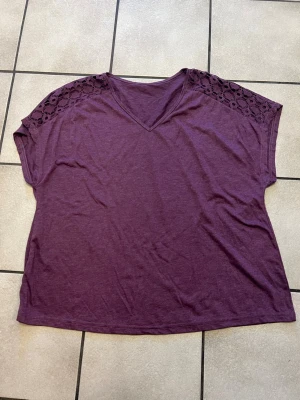 Lila topp med spetsdetaljer storlek XL/XXL - Lila topp med v-ringning och snygga spetsdetaljer på axlarna. Modellen är loose fit och har korta ärmar. Perfekt för dig som gillar en avslappnad och trendig stil. Materialet känns mjukt och bekvämt mot huden.