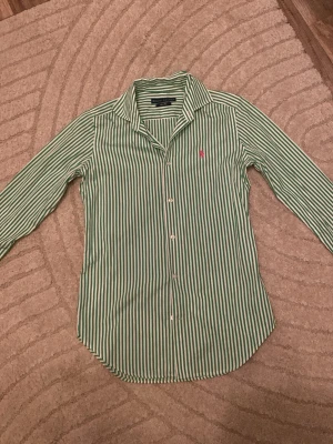 Grön- och vitrandig skjorta Ralph Lauren - Klassisk grön- och vitrandig skjorta från Ralph Lauren med lång ärm och knappar framtill. Skjortan har en broderad rosa logga på bröstet och är tillverkad i mjuk bomull. Perfekt för en stilren och avslappnad look.