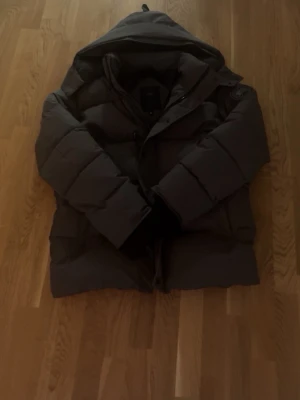 Mörkgrå dunjacka från Canada Goose - Säljer en mörkgrå dunjacka från Canada Goose med stor huva och puffig design. Jackan har ett emblem på ärmen och praktiska knappar framtill. Perfekt för kalla vinterdagar och riktigt varm tack vare dunfyllningen. Päls ingår i köpet