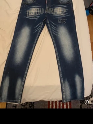 Blå Dsquared2 jeans med tryck - Säljer ett par blå jeans från Dsquared2 med stort tryck på framsidan där det står 'DSQUARED2' och 'Established 1995'. Jeansen har en tvättad look med ljusa partier och klassisk femficksmodell. Perfekta för dig som gillar streetwear och vill sticka ut.
