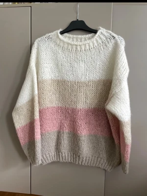 Stickad tröja i pastellfärger NY - Mysig stickad tröja med breda blockränder i vitt, beige, ljusrosa och ljusgrått. Tröjan har rund halsringning, lång ärm och ribbstickade muddar. Perfekt för dig som gillar en oversized och avslappnad look. Materialet känns mjukt och fluffigt.