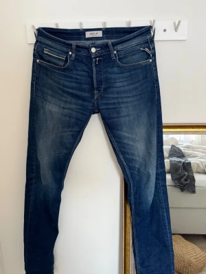 Mörkblå slim fit jeans från Replay  - Snygga mörkblå slim fit jeans från Replay med klassisk femficksmodell och diskreta slitningar framtill. Jeansen har normal midja, dragkedjegylf och detaljerade bakfickor med Replay-logga. Perfekta för en trendig och avslappnad stil. Storlek W30 L34