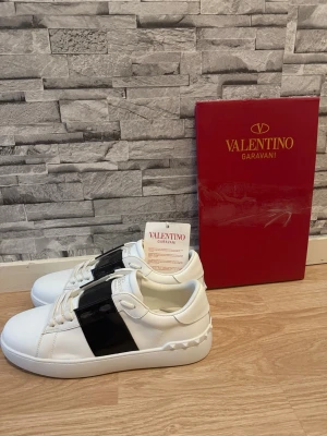 Valentino Garavani vita sneakers - Snygga vita sneakers från Valentino Garavani med en bred svart läderdetalj över mitten. Klassisk låg modell med vita snören och diskret logga på plösen. Tillverkade i slätt läder för en clean och exklusiv look.