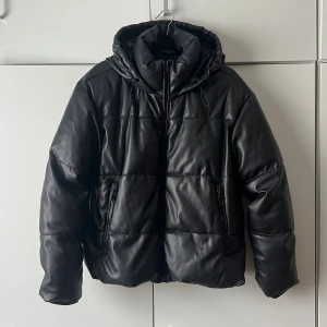 Svart vinterjacka från Zara - Säljer en svart jacka med huva och dragkedja framtill. Jackan har quiltad design och två fickor med dragkedja. Materialet är skinnimitation som ger en cool look. Perfekt för kalla vinterdagar och riktigt snygg att matcha med jeans.