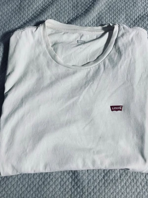 Vit t-shirt från Levi's med logga - Klassisk vit t-shirt från Levi's med rund halsringning och den ikoniska röda Levi's-loggan broderad på bröstet. Tillverkad i mjuk bomull för en skön känsla och enkel stil. Perfekt basplagg för alla tillfällen.