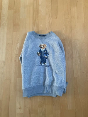 Grå sweatshirt Polo Bear Ralph Lauren - Säljer en grå sweatshirt från Polo Ralph Lauren med den ikoniska Polo Bear-björnen på bröstet, klädd i blå kostym och slips. Tröjan har rund halsringning, ribbade muddar och är tillverkad i mjukt bomullsmaterial. Perfekt för en avslappnad men ändå stilren look. Storlek S
