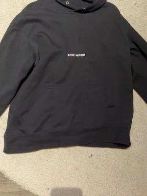 Svart hoodie från Saint Laurent - Snygg svart hoodie från Saint Laurent med minimalistisk logga på bröstet. Klassisk passform med huva och känguruficka framtill. Tillverkad i mjukt material som känns skönt mot huden. Perfekt för dig som gillar stilrena designer och exklusiva märken. Snörena är borta, klippte även av lite information där nere för de kliande material.