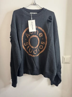 Mörkgrå sweatshirt Acne Studios - Cool mörkgrå sweatshirt från Acne Studios med stort orange tryck framtill. Oversized passform och distressed detaljer med slitningar på axel och ärm. Tillverkad i mjuk bomullsmix och har långa ärmar samt ribbade muddar.