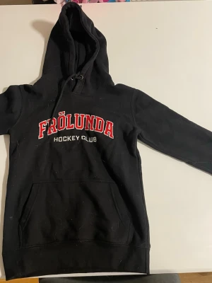Svart Frölunda Hockey Club hoodie - Snygg svart hoodie med stor huva och känguruficka framtill. Frölunda Hockey Club-logga i rött och vitt broderat på bröstet. Klassisk modell med ribbade muddar och dragsko i huvan. Perfekt för dig som vill visa vilket lag du hejar på.