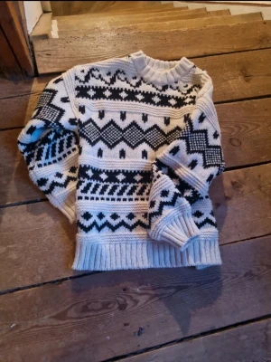 Vit och svart mönstrad stickad tröja - Stickad tröja i vitt med svart grafiskt mönster. Tröjan har rund halsringning, ribbstickade muddar och nederkant. Perfekt för kalla dagar och har en klassisk nordisk vibe. Materialet känns mjukt och varmt, troligen ullblandning.