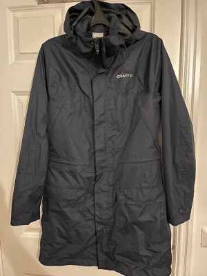 Blå  regnjacka från Craft med huva - Lång blå  regnjacka från Craft med justerbar huva och dragkedja framtill. Jackan har två stora fickor med lock och diskret logga på bröstet. Tillverkad i vattenavvisande material, perfekt för regniga dagar. Enkel och stilren design som passar till det mesta.