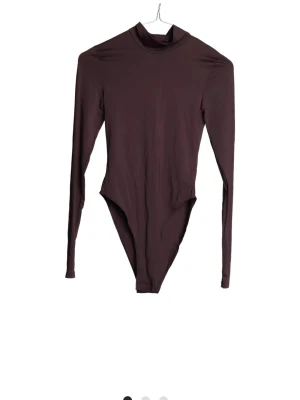 Mörkbrun långärmad body - En stilren mörkbrun body med lång ärm och hög hals. Tillverkad i ett mjukt och stretchigt material som sitter tight mot kroppen. Perfekt att bära under tröjor eller som den är för en clean look.