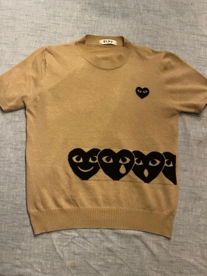 Beige stickad tröja Comme des garcon - Säljer en beige stickad tröja från PLAY med korta ärmar. Tröjan har svarta hjärtan med olika ansiktsuttryck längs nederkanten och ett litet hjärta på bröstet. Rund halsringning och ribbade muddar. Perfekt för dig som gillar unik design.