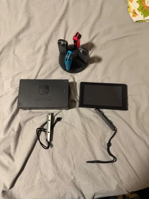 Nintendo Switch konsol med tillbehör - Nintendo Switch konsol med svart docka, Joy-Con-kontroller i rött, blått och svart, laddningsstation för kontroller samt kablar. Skärmen är svart och rektangulär, och tillbehören är i plast. Perfekt för gaming och multiplayer.