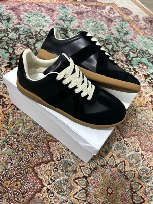 Svarta Maison Margiela Replica sneakers - Maison Margiela Replica sneakers i svart läder och mocka med beige sula och vita snören. Klassisk låg modell med retrovibbar och diskret logga på plösen. Perfekta för dig som gillar stilrena och tidlösa sneakers.