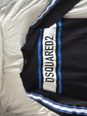 Svart sweatshirt från Dsquared2 - Svart sweatshirt från Dsquared2 med vita och blå ränder längs ärmarna och ett stort vitt tryck med loggan på bröstet. Tröjan har rund halsringning och ribbade muddar. Perfekt för en sportig och trendig look.