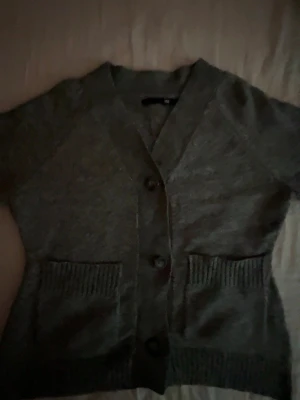 Mörkgrå stickad kofta med knappar - Mörkgrå stickad kofta med v-ringning och stora knappar framtill. Koftan har två ribbstickade fickor och långa ärmar. Perfekt för lager-på-lager och chill dagar. Materialet ser ut att vara mjukt och bekvämt.