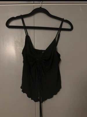 Svart linnetopp med snörning från Ginatricot - Snygg svart linnetopp från Ginatricot med tunna axelband och trendig snörning framtill. Toppen har vågiga kanter nertill och en croppad passform som ger en cool vibe. Perfekt att styla med jeans eller kjol för en modern look.