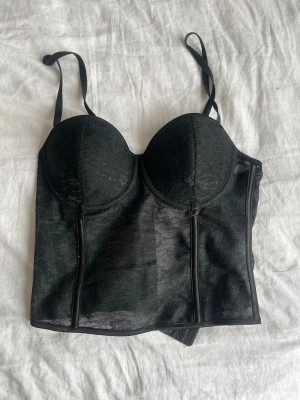 Svart spetskorsett med kupor - Snygg svart korsett med formade kupor och justerbara axelband. Materialet är spets med diskret mönster och genomskinliga partier. Korsetten har markerade sömmar framtill som ger en cool och strukturerad look.