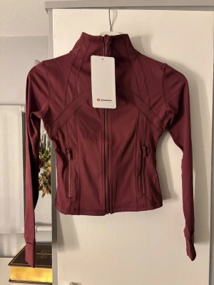 Vinröd träningsjacka från Lululemon - Snygg vinröd träningsjacka från Lululemon med figurnära passform, hög krage och dragkedja framtill. Jackan har långa ärmar med tumhål och två dragkedjefickor. Tillverkad i stretchigt funktionsmaterial som andas. Finns även matchande byxor!!!
