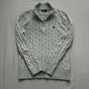 Grå kabelstickad tröja Polo Ralph Lauren - Kabelstickad grå tröja från Polo Ralph Lauren med halv dragkedja och hög krage. Tröjan har ribbstickade muddar och nederkant samt den klassiska broderade loggan på bröstet. Perfekt för lager-på-lager och snygg till jeans.