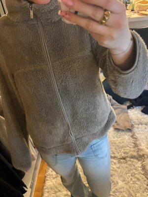 Beige teddyjacka med dragkedja - Mysig beige teddyjacka med hög krage och hel dragkedja framtill. Jackan har en avslappnad passform och är gjord i fluffigt, mjukt material som ger en skön och trendig look. 