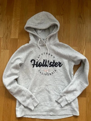  hoodie Hollister - Ljusgrå hoodie från Hollister med broderad logga och texten 'Southern California' på bröstet. Klassisk känguruficka framtill, justerbar dragsko i huvan och ribbade muddar. Mjuk bomullsblandning som känns skön mot huden. Perfekt för en avslappnad och trendig look.