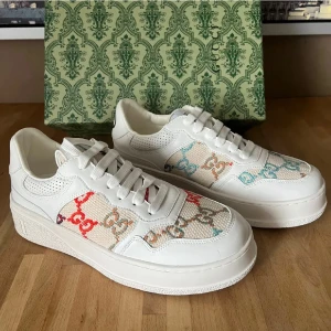 Gucci vita sneakers med färgglada detaljer - Snygga vita sneakers från Gucci med färgglada GG-loggor på sidan och guldigt GG-tryck på hälen. Skorna har en låg profil, perforerade detaljer och är tillverkade i läder och canvas. Perfekt för dig som vill sticka ut med exklusiva skor.