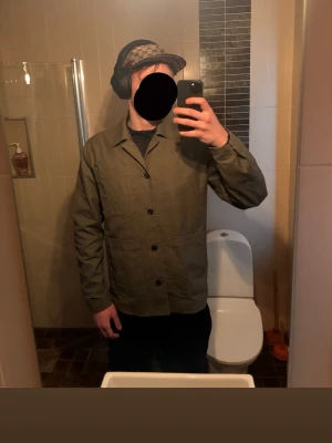 Olivgrön overshirt med knappar - Stilren olivgrön overshirt med klassisk krage och tre framfickor. Modellen har raka ärmar och stängs med svarta knappar framtill. Materialet ser ut att vara bomull eller en bomullsblandning, perfekt för lager-på-lager. Enkel och clean design som passar till det mesta.