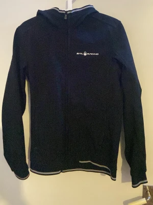 Svart hoodie från Sail Racing - Säljer en svart jacka från Sail Racing med dragkedja framtill och vit logga på bröstet. Hoodien har huva och vita detaljer längs kanterna samt en logga på huvan. Perfekt för dig som gillar stilren och sportig design.