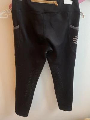 Svarta Ridbyxor - Säljer ett par svarta rid Byxor med gråa detaljer och fickor på sidorna. De har en guldfärgad dragkedja bak och mönstrade partier på benen för extra stil. Perfekta för en sportig look eller till vardagsoutfiten.