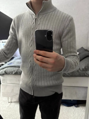 Ljusgrå ribbad ziptröja maison margiella - Säljer en ljusgrå ziptröja med ribbad struktur och diskret kabelstickat mönster framtill. Tröjan har hög krage, dragkedja hela vägen och breda muddar vid ärmslut och nederkant. Perfekt för lager på lager och enkel att matcha.