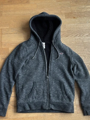 Mörkgrå hoodie från Hollister - Mörkgrå hoodie från Hollister i storlek S med dragkedja, två fickor framtill och mjukt teddyfoder i huvan. Diskret logga på bröstet och dragsko i huvan. Hör av dig vid frågor!
