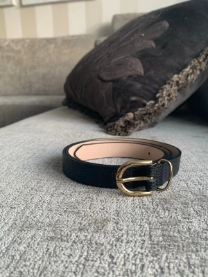 Svart smalt bälte med guldfärgat spänne - Snyggt och stilrent svart bälte i skinn (veganskt-konstläder) med guldfärgat, rundat spänne. Bältet är smalt och passar perfekt till både jeans och kjol. Enkel design som ger en clean look till din outfit.