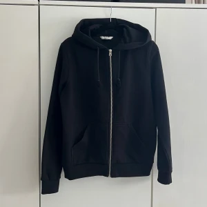 Svart hoodie med dragkedja - Klassisk svart hoodie med dragkedja framtill och två fickor. Har justerbar huva med snören och ribbade muddar vid ärmar och nederkant. Perfekt basplagg för en avslappnad stil. Materialet känns mjukt och bekvämt, troligen bomullsblandning.