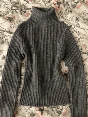 Grå ribbstickad tröja  - Mysig grå ribbstickad tröja med lång ärm och hög krage. Tröjan har en figurnära passform och är tillverkad i ett mjukt, tjockt material som passar perfekt för kyliga dagar. Enkel och stilren design som är lätt att matcha.