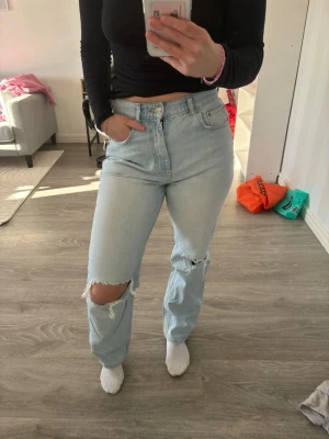 Ljusblå ripped jeans - Säljer ett par ljusblå mom jeans med hög midja och raka ben. Jeansen har slitningar och hål på knäna för en trendig, avslappnad look. Klassisk femficksmodell och tillverkade i denim. Perfekta för dig som gillar en casual och cool stil.