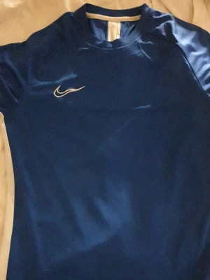 Blå Nike sporttröja med vita detaljer - Säljer en blå sporttröja från Nike med vita sidopaneler och klassisk Nike-logga på bröstet. Tröjan har korta ärmar och är tillverkad i ett lätt, funktionsmaterial som passar perfekt till träning eller match. Strlk M, passar s oxå. Pyttelite hål i nacken som man knappt märker