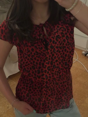 Röd leopardmönstrad blus H&M - Snygg röd blus från H&M med svart leopardmönster. Blusen har korta ärmar, rund halsringning med knytband och är tillverkad i ett tunt, lätt material som känns luftigt. 