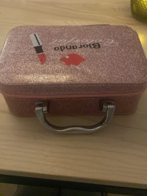 Rosa glittrig sminkväska Floranda - Snygg rosa sminkväska från Floranda med glittereffekt, dekorerad med ett läppstift och röda läppar på locket. Väskan har ett praktiskt handtag i metall och dragkedja runt om. Perfekt för att förvara smink eller skönhetsprodukter.Sminket är med om man vill men kostar extra då  om man vill bara har väskan är et okej ❤️Lådan kostade ny 200 kr 