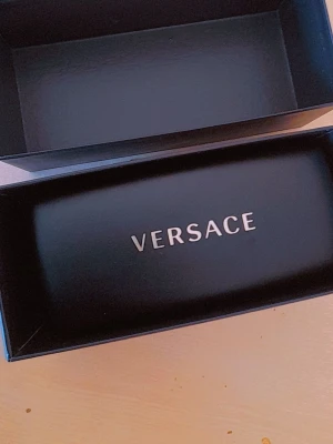 Versace svarta solglasögon med guld - Snygga svarta solglasögon från Versace med ikoniska guldfärgade Medusa-detaljer på skalmarna. Glasögonen har en klassisk rektangulär form och levereras med originaletui. Perfekt accessoar för dig som vill sticka ut.