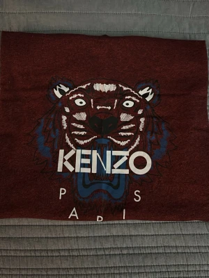 Röd Kenzo t-shirt med tigertryck - Snygg röd t-shirt från Kenzo med det ikoniska tigertrycket och texten 'KENZO PARIS' på bröstet. T-shirten har rund hals och korta ärmar. Perfekt för dig som vill sticka ut med en trendig och ungdomlig stil.