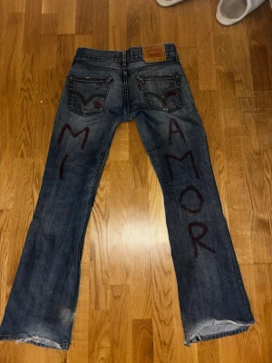 Levi's jeans med text MI AMOR - Unika blå Levi's jeans med handmålad text 'MI AMOR' på baksidan av benen. Klassisk femficksmodell i slitstark denim med bootcut/utsvängd passform och läderpatch bak i midjan. Perfekta för dig som vill sticka ut med en personlig touch.