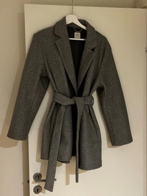 Grå kappa med knytskärp från ONLY - Stilren grå kappa från ONLY med klassisk krage och knytskärp i midjan. Kappan har raka ärmar, två sidofickor och är tillverkad i ett mjukt tyg. Skriv vid frågor!