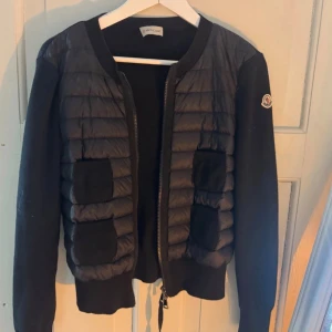 Svart cardiganjacka från Moncler - Snygg svart cardiganjacka från Moncler med quiltad framsida och två fickor. Jackan har dragkedja, ribbade muddar och Moncler-logga på ärmen. Materialmix med vadderad front och stickade ärmar ger en modern look.