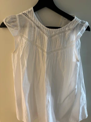 Vit blus med spetsdetaljer - Söt vit blus med spetsdetaljer och veck framtill. Blusen har en luftig passform, rundad halsringning och är ärmlös. Perfekt till sommaren och enkel att matcha med jeans eller kjol. Materialet känns lätt och andas bra. Köpte blusen två år sedan i Spanien på Mango Kids, endast använd en gång därav väldigt bra skick!