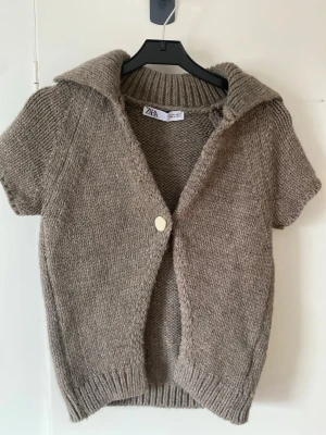 Beige stickad kortärmad kofta från Zara - Stickad beige kofta från Zara med korta ärmar, bred krage och en vit knapp framtill. Koftan har ribbstickade detaljer vid krage, ärmslut och nederkant. Perfekt att slänga över en topp för en avslappnad look och aldrig använd💗