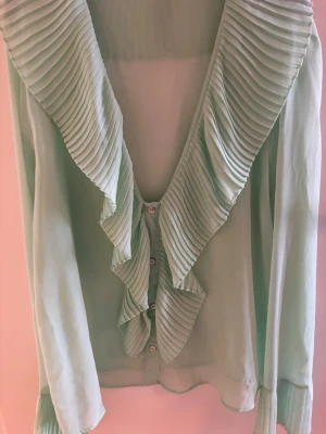 Mintgrön blus med volangkrage - Superfin mintgrön blus med stora plisserade volanger längs kragen och framtill. Blusen har knäppning framtill och långa ärmar med plisserade detaljer vid ärmsluten. Tunt, lätt material som ger en luftig känsla.