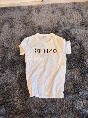 Vit Kenzo t-shirt med färgglad logga - Säljer en vit t-shirt från Kenzo med färgglad logotyp framtill. T-shirten har klassisk passform, rund halsringning och korta ärmar. Tillverkad i mjuk bomull som känns skön mot huden. Perfekt för dig som gillar stilrena plagg med en twist.