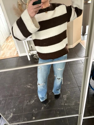 Brunvit randig stickad tröja - Säljer en oversized stickad tröja med breda bruna och vita ränder. Tröjan har lång ärm, rund halsringning och är gjord i ett mjukt, fluffigt material som känns riktigt skönt mot huden. Perfekt för dig som gillar en avslappnad och trendig stil. Från Gina 14+ i storlek M, men passar mig som har S. 